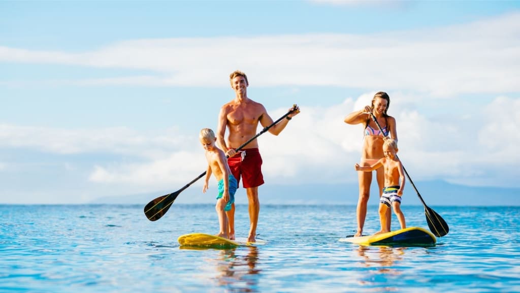 Two adults and two children paddleboarding on calm water under a blue sky, each holding a paddle and standing on their own board.